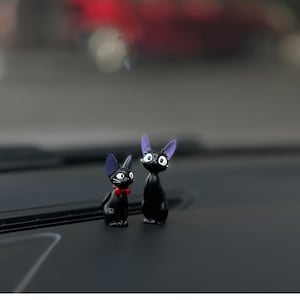May include: Two black cat figurines with purple ears and white eyes. The cat on the left has a red bow tie. The cats are sitting on a black surface.