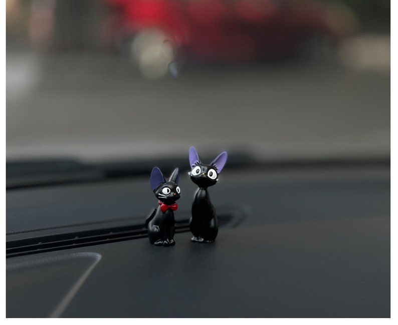 May include: Two black cat figurines with purple ears and white eyes. The cat on the left has a red bow tie. The cats are sitting on a black surface.