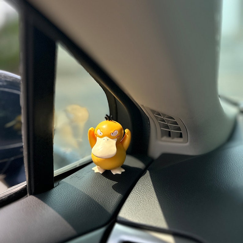 May include: A yellow duck-like Pokemon figurine with angry eyes and a black tuft of hair. The figurine is sitting on the dashboard of a car.