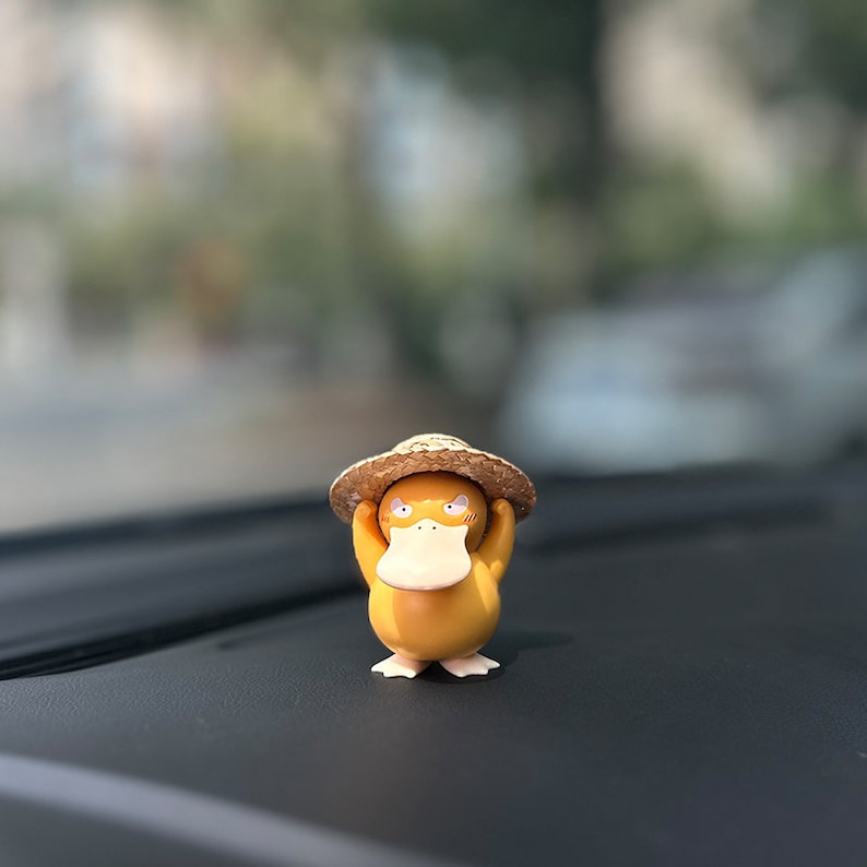 May include: A yellow duck figurine wearing a brown straw hat. The duck is holding the hat with its hands and has a surprised expression on its face. The figurine is sitting on a black surface.