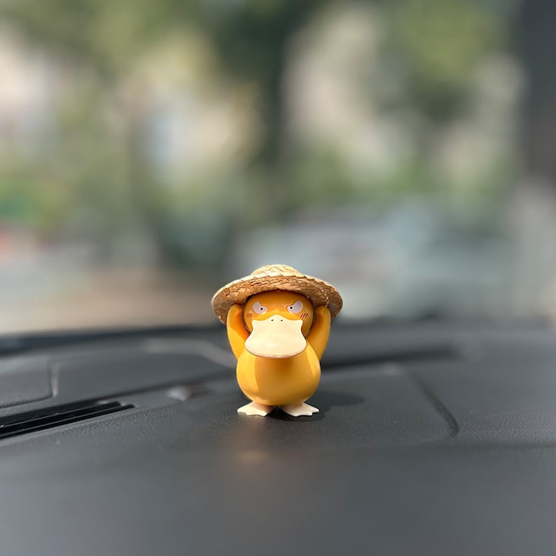 May include: A yellow duck figurine wearing a brown straw hat. The duck has a grumpy expression and is holding the hat with its hands.