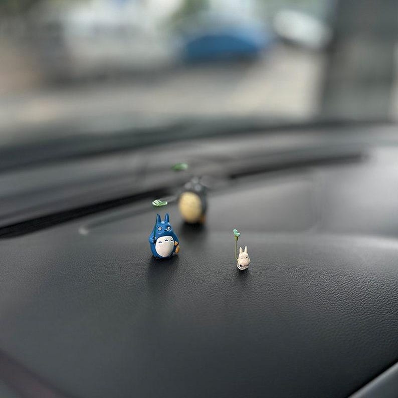 May include: Three small figurines of characters from the animated film 'My Neighbor Totoro' are displayed on a black surface. The figurines are made of a light blue, white, and gray material. The figurines are about 2 inches tall.