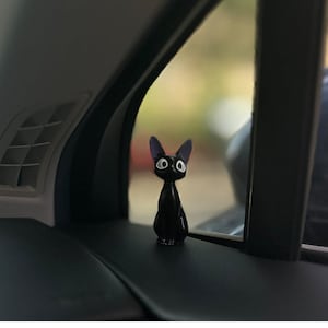 May include: Black cat figurine with large eyes, sitting on a car dashboard.