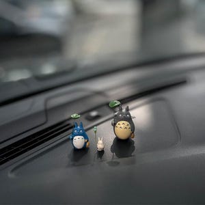 May include: Three small figurines of characters from the animated film 'My Neighbor Totoro'. The figurines are made of painted resin and feature Totoro, a small Totoro, and a cat-like creature. The figurines are arranged on a black surface.