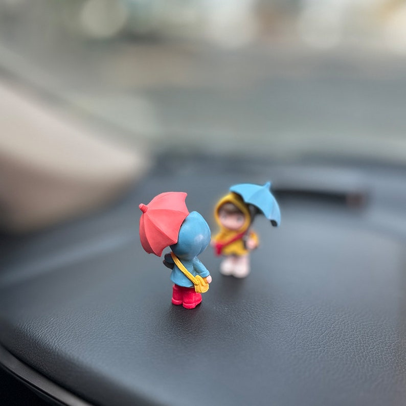 May include: Two small figurines of people holding umbrellas. The person on the left is wearing a blue raincoat and holding a red umbrella. The person on the right is wearing a yellow raincoat and holding a blue umbrella.