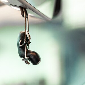 May include: A black and white character figurine hanging from a rearview mirror. The character is sitting on a swing and has a white face mask.