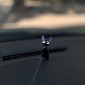 May include: A black cat figurine with purple ears and white eyes. The figurine is sitting on a black surface.