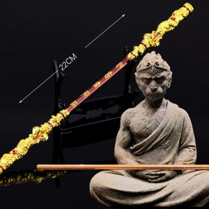 K&ouml;nnte beinhalten: Ein goldener und roter Stab mit chinesischen Schriftzeichen, 56 cm lang, wird neben einer Steinstatue eines Affen in sitzender Meditationshaltung gezeigt.