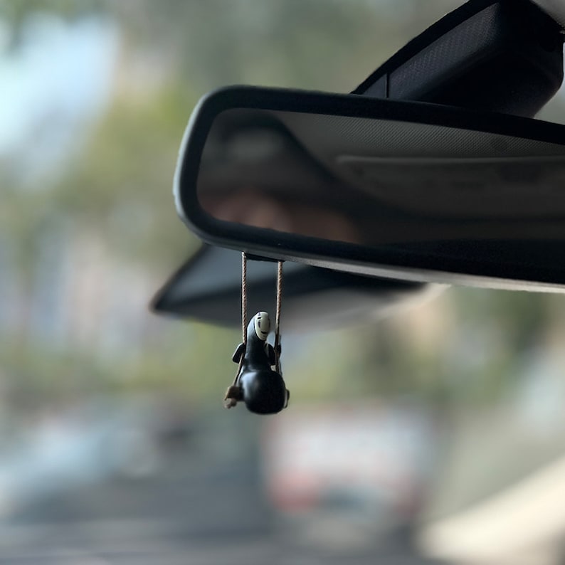 May include: A black and white character figurine hanging from the rearview mirror of a car. The character is from the animated film Spirited Away.