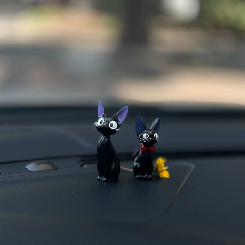 May include: Two black cat figurines with large eyes and purple ears. The cat on the left is looking to the right, while the cat on the right is looking straight ahead. The cat on the right is wearing a red bow tie. The figurines are sitting on a black surface.