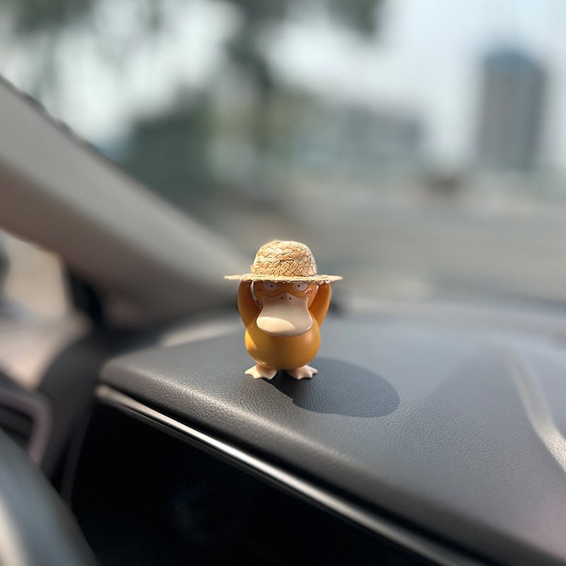 May include: A yellow duck figurine wearing a straw hat, sitting on a car dashboard.