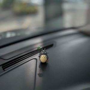 May include: A small, black and yellow figurine of a character from the animated film My Neighbor Totoro, holding a small green leaf. The figurine is sitting on the dashboard of a car.