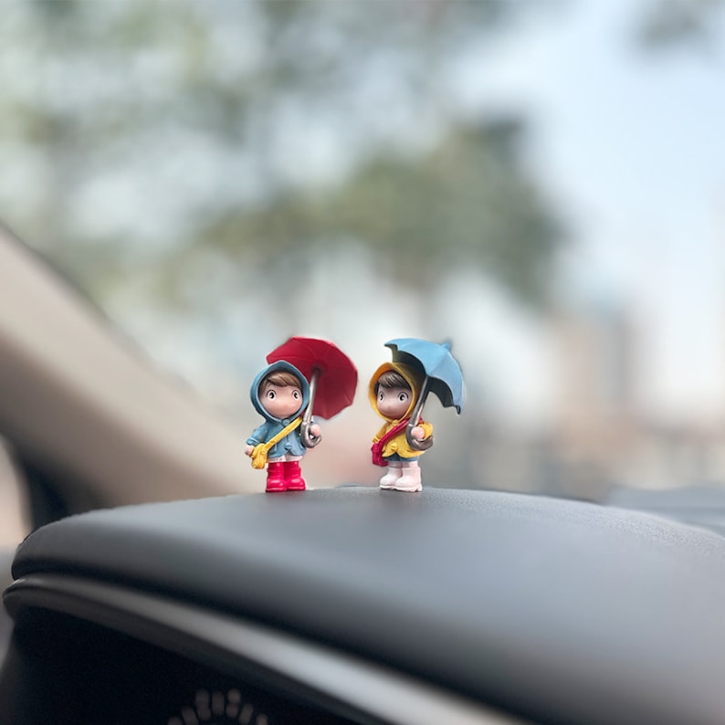 May include: Two small figurines of children wearing raincoats and holding umbrellas. The child on the left is holding a red umbrella and the child on the right is holding a blue umbrella. The figurines are sitting on a car dashboard.