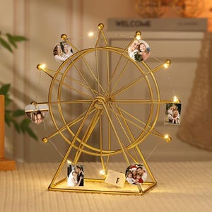 May include: A gold-colored Ferris wheel photo display with small, illuminated lights. The display holds several small photos. The base is a geometric shape. The background includes a wooden frame and a sign that says "WELCOME".