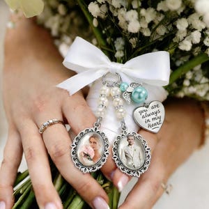 Może przedstawiać: Bukiet ślubny z białych kwiatów i białą wstążką. Dwie owalne zawieszki ze zdjęciami i ozdobnymi srebrnymi ramkami wiszą na wstążce. Jedna zawieszka przedstawia kobietę z różowymi kwiatami, druga mężczyznę w garniturze.