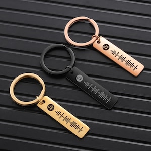 Op de afbeelding: Drie sleutelhangers met verschillende metalen afwerkingen: goud, zwart en rosé goud. Elke sleutelhanger heeft een klein muzieknotensymbool en een barcode-achtig ontwerp.