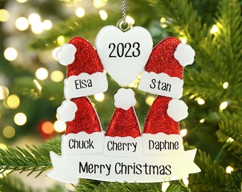 May include: White Christmas ornament with a red glitter heart in the center that says "2023".  Four red glitter Santa hats surround the heart with names "Elsa", "Stan", "Chuck", "Cherry", and "Daphne" written below.  A white banner at the bottom says "Merry Christmas".