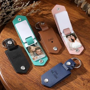 May include: A collection of personalized leather keychains in various colors: black, teal, brown, pink, and blue. Each keychain features a metal ring and a snap closure. Some keychains display the word "Dad" or "Dada" and include a photo.