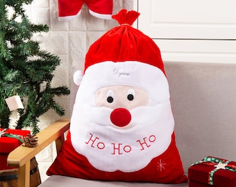 May include: A large red Santa Claus gift bag with a white beard and face, embroidered with "HO HO HO". The bag has a drawstring closure and the name "Dylan" embroidered on the white beard. A matching red stocking with the name "Barrell" is hanging in the background.