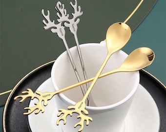May include: A collection of decorative spoons and stirrers. Two gold spoons with rounded bowls and two silver stirrers with antler designs are visible. The spoons and stirrers are resting in a white teacup and saucer.