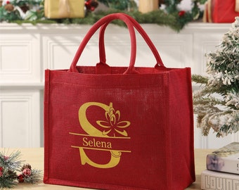 Könnte beinhalten: Eine rote Jute-Tragetasche mit zwei Griffen. Die Tasche hat einen goldfarbenen Buchstaben "S" mit floralem Design und den Namen "Selena" darunter. Die Tasche steht auf einer Holzoberfläche.
