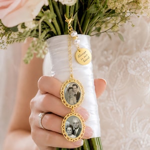 Op de afbeelding: Een goudkleurige trouwboeketbedel met twee ovale frames met zwart-witfoto's, een kleine ronde hanger met de inscriptie Always in our heart en twee witte parelkralen bevestigd aan een gouden ketting met een karabijnsluiting.