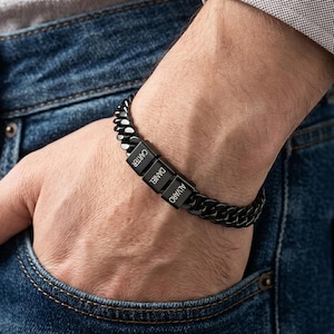 May include: A black metal curb chain bracelet featuring three rectangular beads engraved with the names Carter, Daniel, and Alvaro. The bracelet has a polished finish and a sturdy link design that wraps around the wrist, showcasing a dark aesthetic.