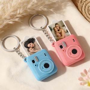May include: Two camera-shaped keychains, one blue and one pink, each holding a small photo. The keychains have a silver chain and ring. The photos show a mother and child. The cameras have a lens and flash.