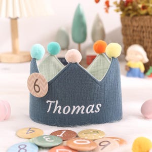 May include: A blue fabric crown with colorful pom-poms and the name "Thomas" embroidered on the front. The crown has a number "6" on the side.