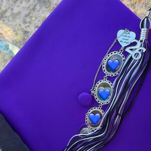 K&ouml;nnte beinhalten: Eine leuchtend violette Doktorhut-Kopfbedeckung mit einer mehrschichtigen Quaste in Violett, Schwarz und Wei&szlig;. An der Quaste befinden sich ein silberner Anh&auml;nger mit dem Text Walk with me across the stage, eine Zahl 26 und drei ovale Fotorahmen.