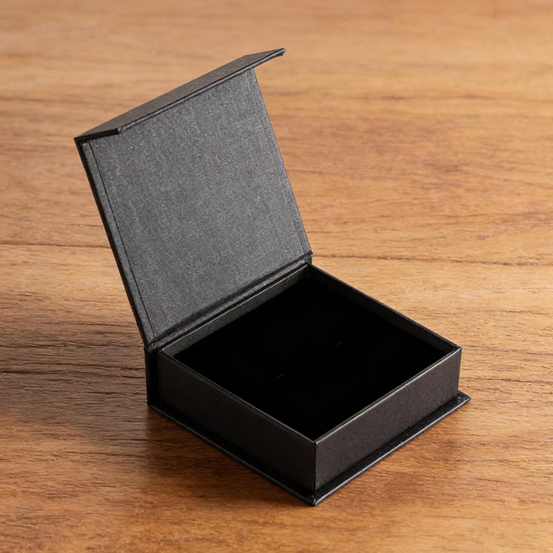 May include: A square, black jewelry box with a hinged lid that is open. The exterior of the box has a textured, matte finish, while the interior base is lined with a smooth, deep black material designed to hold small items securely in place.