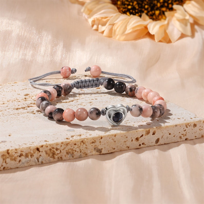 Puede incluir: Un brazalete de cuentas rosa y gris con un dije de coraz&oacute;n plateado. El dije de coraz&oacute;n tiene una peque&ntilde;a ventana redonda y transparente en el centro.