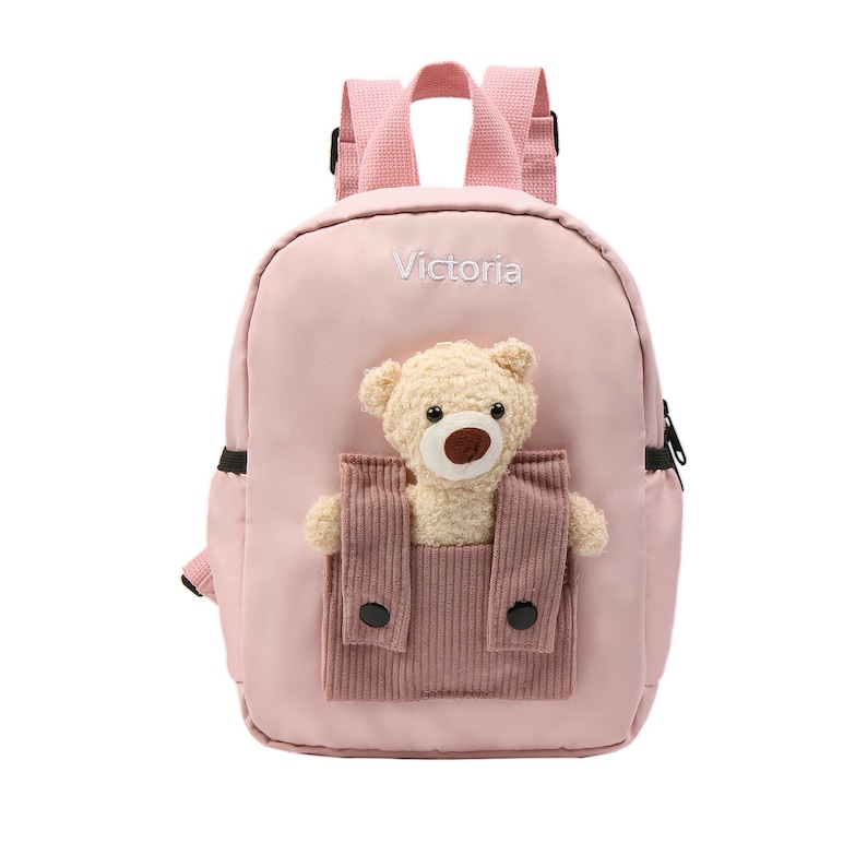 May include: A light pink backpack featuring the name Victoria embroidered in white thread. The front includes a textured corduroy pocket containing a plush beige teddy bear with a brown nose and black eyes, complemented by matching pink straps.