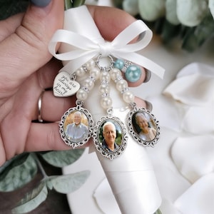 May include: A wedding bouquet charm featuring three oval silver-toned frames with portraits, a heart-shaped pendant engraved with the text 'you are always in my heart', white pearl beads, and a light blue bead, all tied with a white satin ribbon bow.