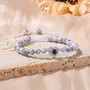 Puede incluir: Pulsera de cuentas blancas con un dije de coraz&oacute;n plateado que tiene una peque&ntilde;a imagen redonda negra y plateada en su interior. La pulsera es ajustable con un cord&oacute;n blanco.