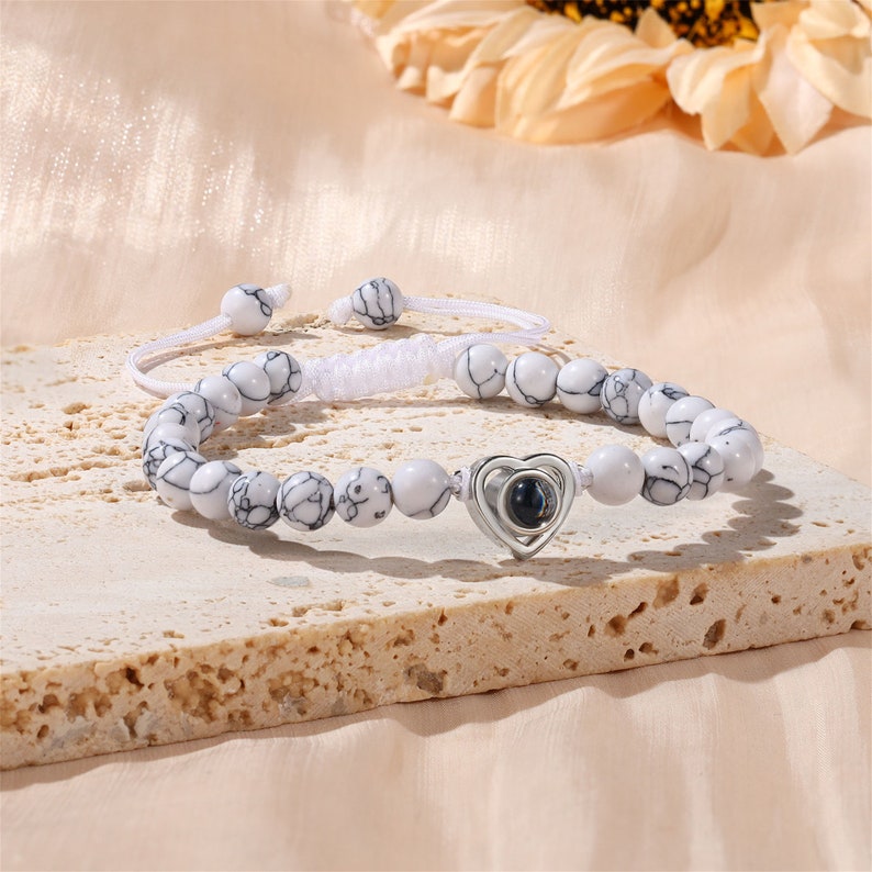 Puede incluir: Pulsera de cuentas blancas con un dije de coraz&oacute;n plateado que tiene una peque&ntilde;a imagen redonda negra y plateada en su interior. La pulsera es ajustable con un cord&oacute;n blanco.