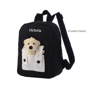 May include: A black backpack featuring a white corduroy front pocket with a plush beige teddy bear peeking out. The name Victoria is embroidered in white text above the pocket, and a line points to the name with the text Custom Name.