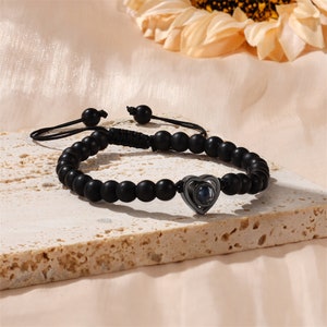 Puede incluir: Un brazalete de cuentas negras con un dije de coraz&oacute;n plateado que tiene una peque&ntilde;a lente en el centro. El brazalete est&aacute; sobre una superficie texturizada gris claro.