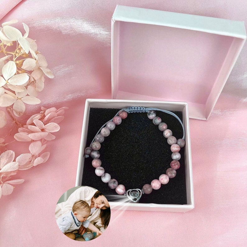 Puede incluir: Un brazalete de cuentas rosa y gris con un dije de coraz&oacute;n plateado en una caja de regalo blanca. La caja est&aacute; abierta y el brazalete est&aacute; dentro. El brazalete est&aacute; sobre un fondo negro.