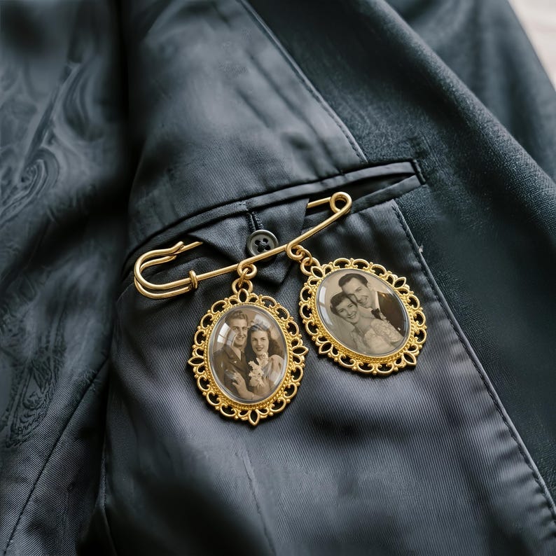 May include: A gold-toned metal safety pin brooch featuring two oval-shaped pendants with ornate filigree frames. Each pendant contains a sepia-toned photograph of a couple. The brooch is pinned to the dark fabric of a suit jacket interior.