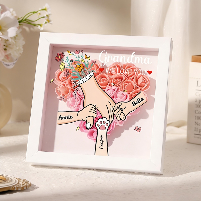 May include: A white framed artwork with a heart-shaped floral arrangement in shades of pink and peach. The artwork features illustrations of hands holding each other, with the names "Annie", "Bella", and "Cooper" inscribed. The text "Grandma WE LOVE YOU" is also present.