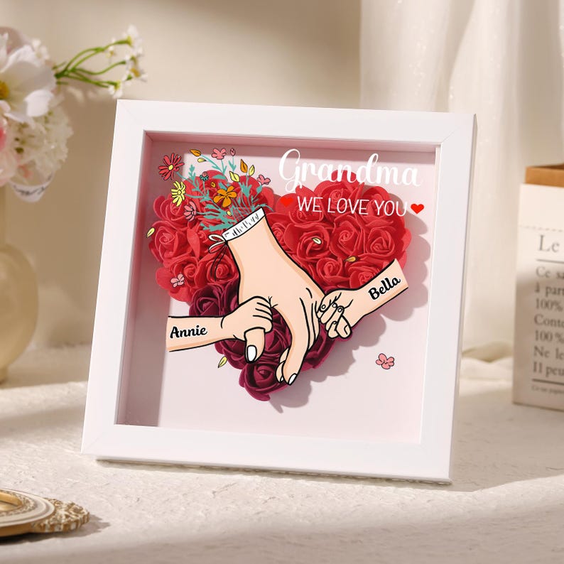 May include: A framed artwork featuring a heart-shaped arrangement of red roses. The artwork includes illustrations of hands holding each other, with the names "Annie" and "Bella" on the hands. The text "Grandma WE LOVE YOU" is displayed above the heart.