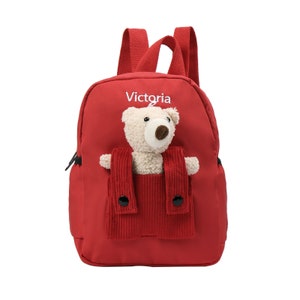 May include: A bright red backpack featuring the name Victoria printed in white above a front pocket. A plush cream-colored teddy bear is tucked into the pocket, which has two red corduroy straps with black buttons attached to the front.