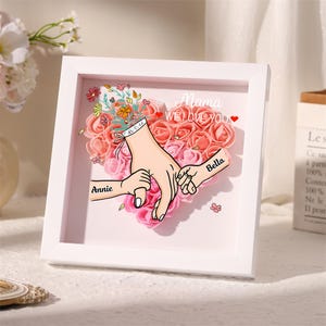 May include: White framed artwork with a heart of pink roses. The artwork shows illustrations of hands holding each other, with the names "Annie" and "Bella" on the hands. The text "Mama WE LOVE YOU" is also present.