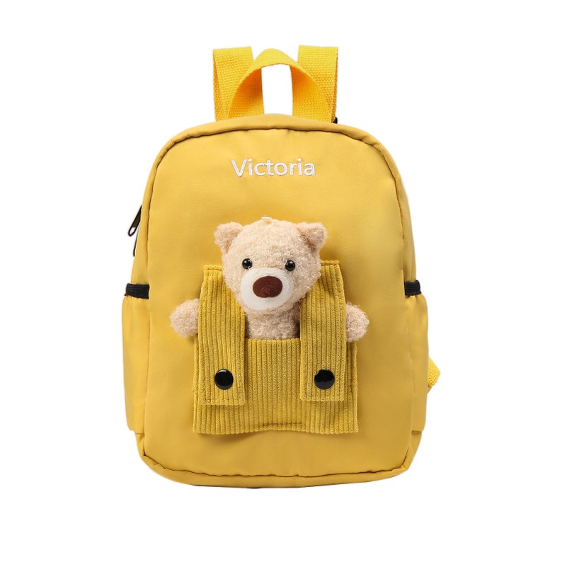May include: A bright yellow backpack featuring the name Victoria embroidered in white text. The front includes a plush beige teddy bear peeking out from a corduroy pocket with two black buttons. The bag has yellow shoulder straps and side pockets.