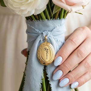May include: A gold oval locket featuring an engraved black line drawing of a flower and the name Sabrina, suspended from a light blue frayed ribbon tied around a bouquet of white and pale pink roses and ranunculus flowers.