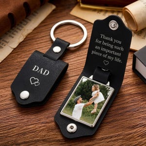 May include: A black leather keychain with a silver metal ring and a snap closure. The exterior is engraved with the word DAD and two hearts. The interior features a rectangular photo frame displaying a person lifting a child and text reading Thank you for being such an important piece of my life with a heart symbol.