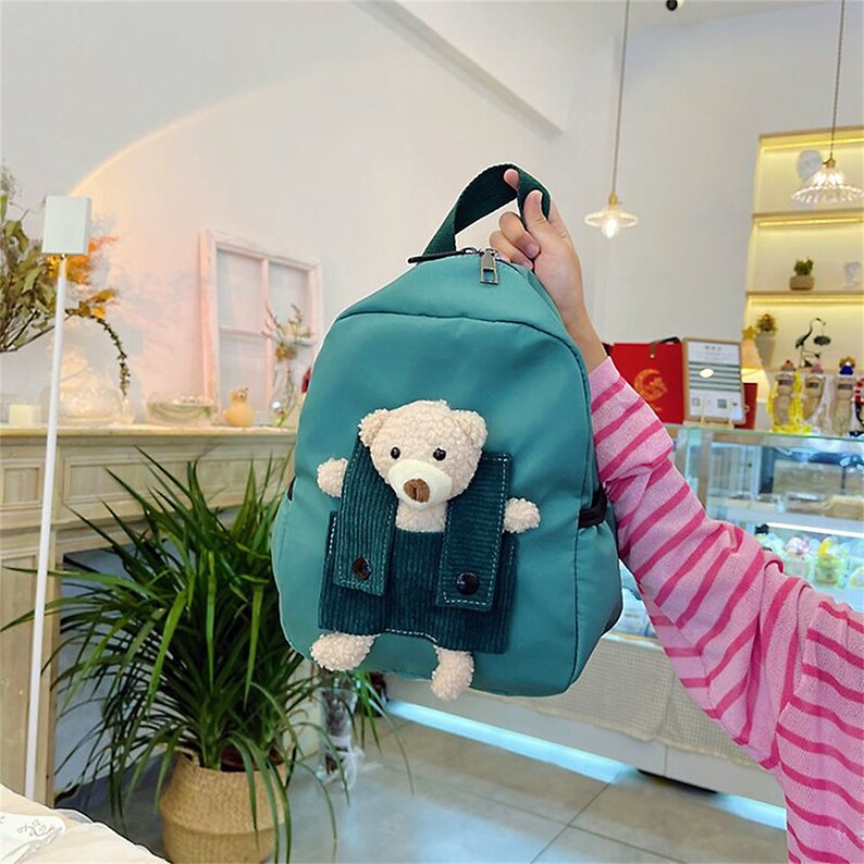 May include: A teal backpack featuring a plush beige teddy bear attached to the front. The bear is positioned inside a dark green corduroy pocket with two black buttons. The backpack has a top handle and a silver zipper closure at the top.