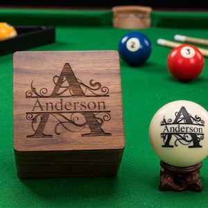 May include: A square wooden box with a dark brown grain featuring a laser-engraved letter A and the name Anderson, accompanied by a white billiard ball displaying the same black letter A and Anderson text, resting on a small dark wooden stand.