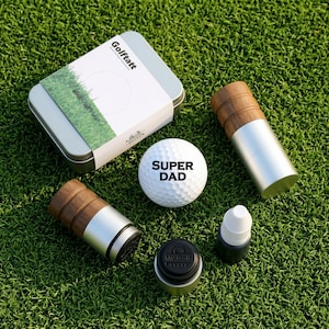 May include: A golf ball stamp kit featuring a white golf ball printed with the text SUPER DAD, a silver and wood-toned stamp tool, a small black ink bottle, a black stamp cap with the text My Golf, and a rectangular metal storage tin.